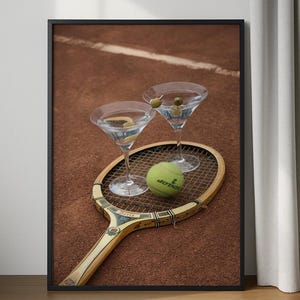 May include: Framed print featuring two martini glasses with olives, a tennis ball, and a vintage tennis racket. The background is a clay tennis court. The martini glasses are filled with a clear liquid and garnished with olives.