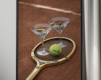 May include: Framed print showing two martini glasses with olives, a tennis ball, and a vintage tennis racket. The background is a clay tennis court. The martini glasses are filled with a clear liquid and garnished with olives.