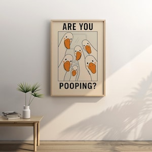 Op de afbeelding: Een ingelijste prent met een cartoon illustratie van witte ganzen met oranje snavels. De tekst "ARE YOU POOPING?" wordt in zwarte hoofdletters weergegeven. Het kunstwerk staat in een lichtgekleurde lijst en hangt aan een beige muur.
