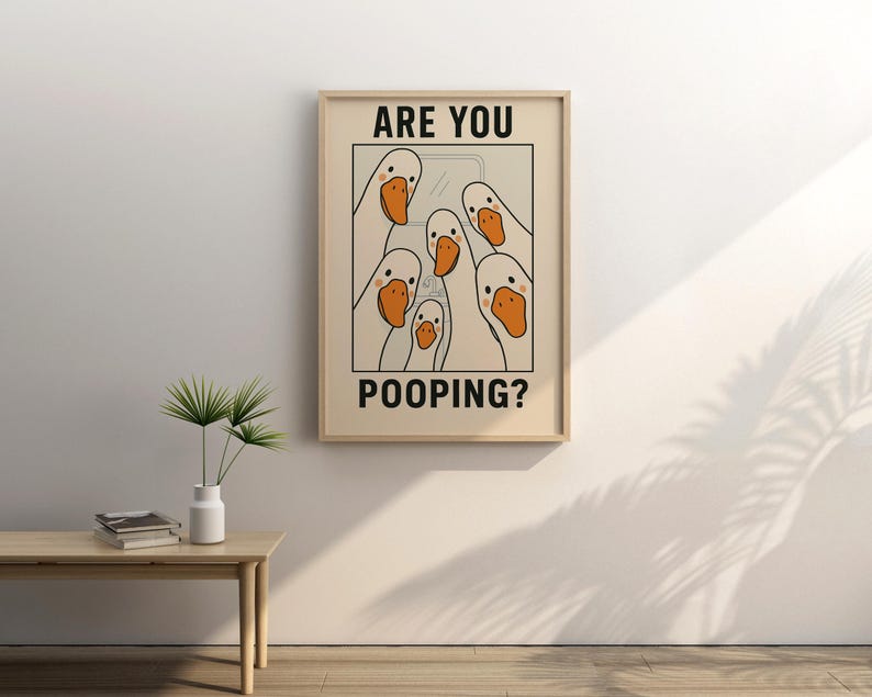 Op de afbeelding: Een ingelijste prent met een cartoon illustratie van witte ganzen met oranje snavels. De tekst "ARE YOU POOPING?" wordt in zwarte hoofdletters weergegeven. Het kunstwerk staat in een lichtgekleurde lijst en hangt aan een beige muur.