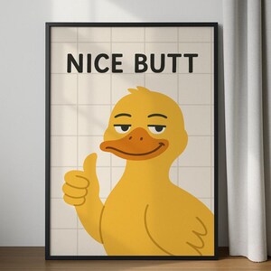 Könnte beinhalten: Gerahmtes Poster mit einer gelben Cartoon-Ente, die einen Daumen hoch zeigt. Die Ente hat einen orangefarbenen Schnabel. Der Text "NICE BUTT" ist in Schwarz gedruckt. Der Hintergrund hat ein Rastermuster. Das Poster ist in einem schwarzen Rahmen.