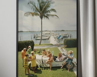 May include: Framed print depicting a retro scene of people in bright clothing around a white convertible. A palm tree and boats are in the background, suggesting a sunny coastal location. The image is set within a black frame.