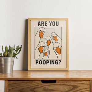 Op de afbeelding: Een ingelijste print met een cartoon illustratie van witte ganzen met oranje snavels. De tekst "ARE YOU POOPING?" staat in zwarte hoofdletters. Het kunstwerk staat in een lichtbeige lijst, geplaatst op een houten oppervlak.