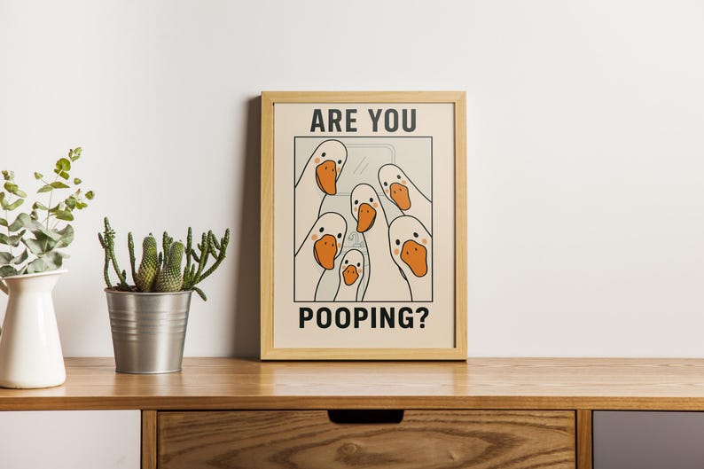 Op de afbeelding: Een ingelijste print met een cartoon illustratie van witte ganzen met oranje snavels. De tekst "ARE YOU POOPING?" staat in zwarte hoofdletters. Het kunstwerk staat in een lichtbeige lijst, geplaatst op een houten oppervlak.