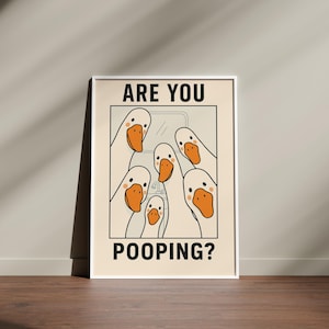 Op de afbeelding: Een ingelijste poster met een cr&egrave;mekleurige achtergrond toont een cartoonillustratie van verschillende witte ganzen met oranje snavels. De tekst "ARE YOU" staat bovenaan en "POOPING?" onderaan, wat een humoristisch ontwerp cre&euml;ert.