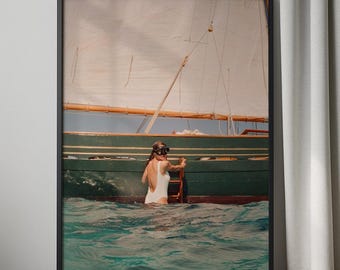 Op de afbeelding: Een ingelijste print met een vrouw in een witte badpak en snorkelmasker die een houten ladder in turkoois water beklimt. Een groene boot en zeil zijn op de achtergrond, met een lichtblauwe lucht. De print zit in een zwart frame.