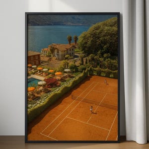 May include: Framed print of a tennis court with players, overlooking a lake and buildings. The court is a reddish-brown color, with a net and white lines. The background features a lake, trees, and buildings with orange umbrellas.