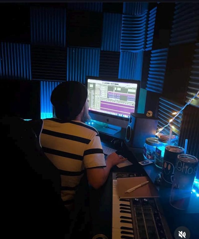 Puede incluir: Una persona con gorro negro y camisa a rayas est&aacute; sentada en un escritorio en un estudio de grabaci&oacute;n. Un monitor de computadora muestra software de edici&oacute;n de audio. Un teclado, altavoces y varios elementos est&aacute;n en el escritorio, iluminados por luz azul.