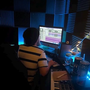 May include: A person wearing a black beanie and a striped shirt is seated at a desk in a recording studio. A computer monitor displays audio editing software. The studio features acoustic foam panels and a keyboard.