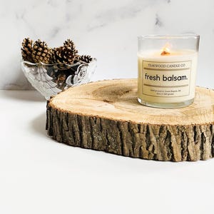 以下が含まれることがあります： Teakwood Candle Co. の「fresh balsam」とラベルされた、木製の芯が付いた透明なガラスのキャンドル。 キャンドルは、松ぼっくりで満たされたガラスのボウルの隣の、素朴な木の輪切りに置かれています。 背景は白い大理石です。
