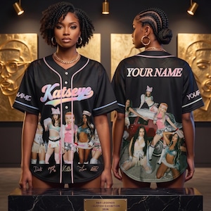 May include: Black baseball jersey with the name "Katseye" in pastel colors across the chest. The jersey features a group of women in various outfits on the front and back. The words "Your Name" are on the back.