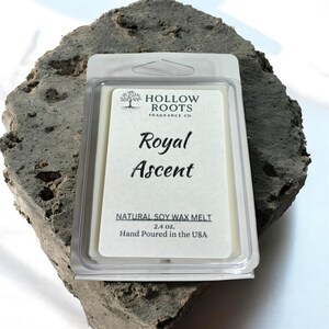 May include: A clear plastic package containing a soy wax melt, labeled "Royal Ascent" by Hollow Roots Fragrance Co. The label is cream-colored with black text and a tree logo. The package is resting on a rough, gray stone.
