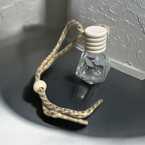May include: A clear, faceted glass bottle with a light wood cap and a beige rope cord. The cord has a wooden bead and yellow accents. The bottle is designed to hold fragrance or essential oils.