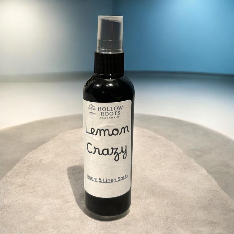 May include: A black spray bottle with a clear cap, labelled "Lemon Crazy Room & Linen Spray" by Hollow Roots Fragrance Co. The bottle is on a light grey surface, with a blue background.