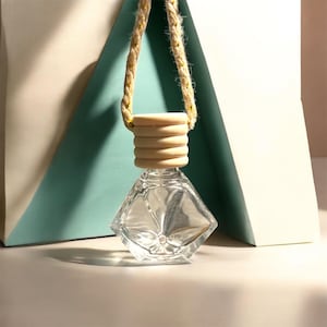 May include: A clear, diamond-shaped glass bottle with a wooden cap and a woven rope for hanging. The bottle is empty and has a decorative design. The background features a light blue and white geometric shape.