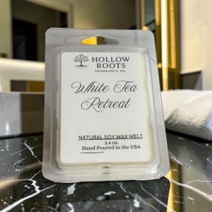 May include: A clear plastic clamshell package containing a white soy wax melt. The label features the text Hollow Roots Fragrance Co., White Tea Retreat, Natural Soy Wax Melt, 2.4 oz., and Hand Poured in the USA, along with a tree icon.