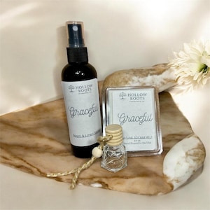 May include: A black spray bottle and a clear glass bottle with a wooden top, both labeled "Graceful." A wax melt and a decorative piece are also present. The items are arranged on a decorative tray.