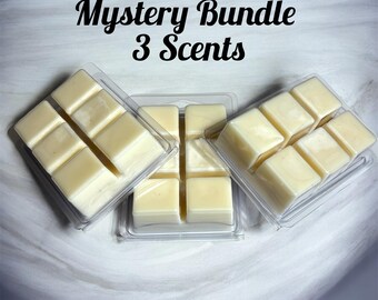 May include: Three clear plastic clamshell containers filled with off-white wax melts, each divided into six individual cubes. The text Mystery Bundle 3 Scents is printed in black script and sans-serif font above the wax melt containers.