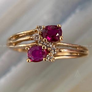 Könnte beinhalten: Ein goldener Ring mit zwei ovalen, tiefroten Rubinen und kleinen Diamanten. Der Ring hat ein geteiltes Schaftdesign und ist in einem goldenen Band gefasst. Die Edelsteine sind zusammengefasst.