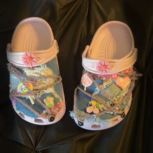 Puede incluir: Un par de zuecos rosa claro personalizados con parches de mezclilla y adornos decorativos. Los zuecos presentan flores rosas, dijes en forma de caramelo y detalles de cuentas. La hora 13:53 est&aacute; visible en la esquina inferior derecha.