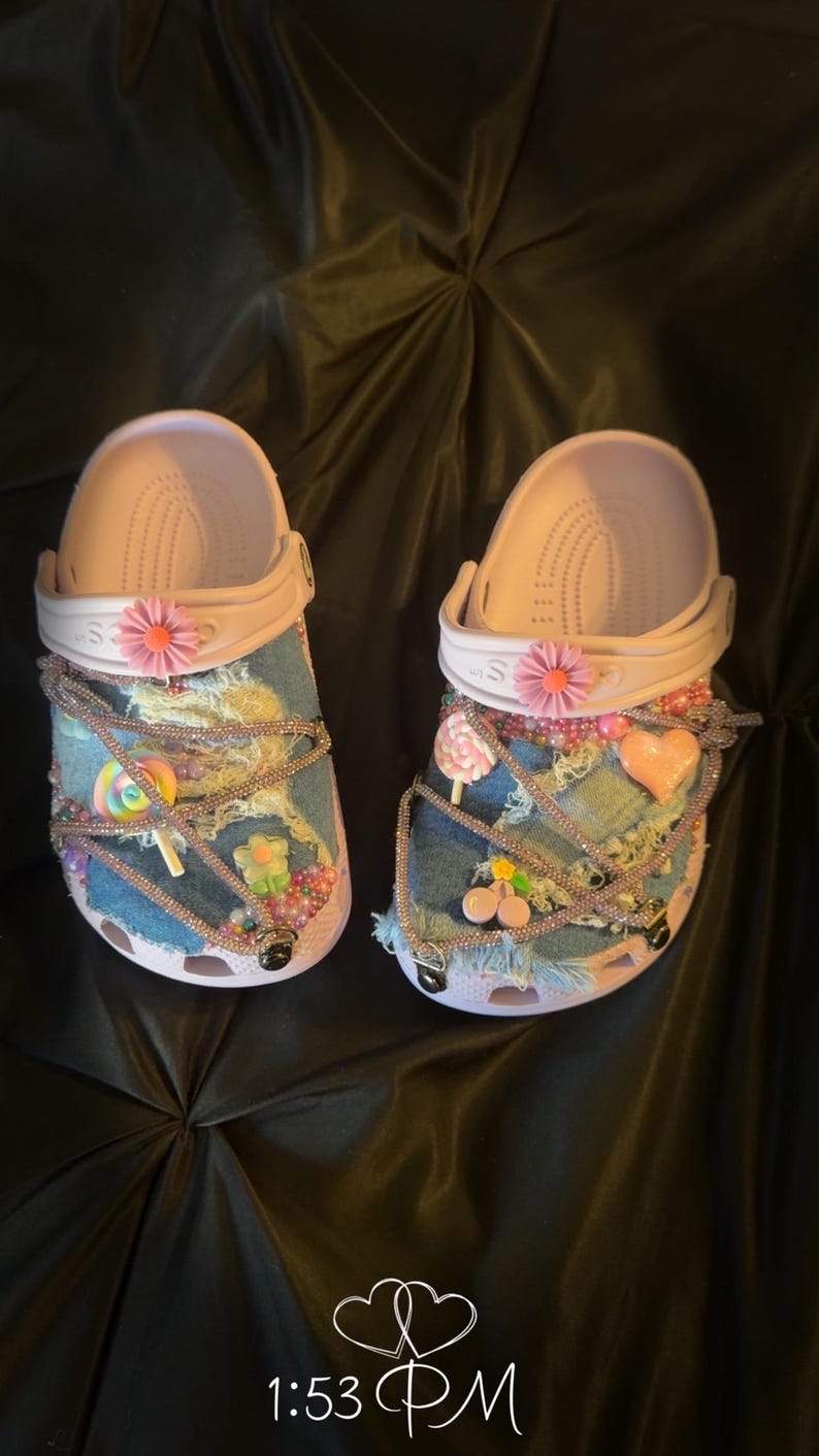 Puede incluir: Un par de zuecos rosa claro personalizados con parches de mezclilla y adornos decorativos. Los zuecos presentan flores rosas, dijes en forma de caramelo y detalles de cuentas. La hora 13:53 est&aacute; visible en la esquina inferior derecha.