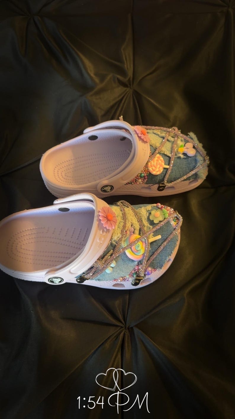 Puede incluir: Un par de zuecos blancos personalizados con parches de mezclilla, pedrer&iacute;a y adornos coloridos. Los zuecos est&aacute;n decorados con flores rosas y naranjas, detalles de cuentas y dijes de piruletas. La hora 13:54 est&aacute; visible en la esquina inferior.