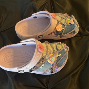 Puede incluir: Un par de zuecos blancos con plantillas de color lila claro, adornados con parches de mezclilla, pedrer&iacute;a y decoraciones florales y de caramelos de colores. Los zuecos tienen una correa y est&aacute;n sobre un fondo negro.