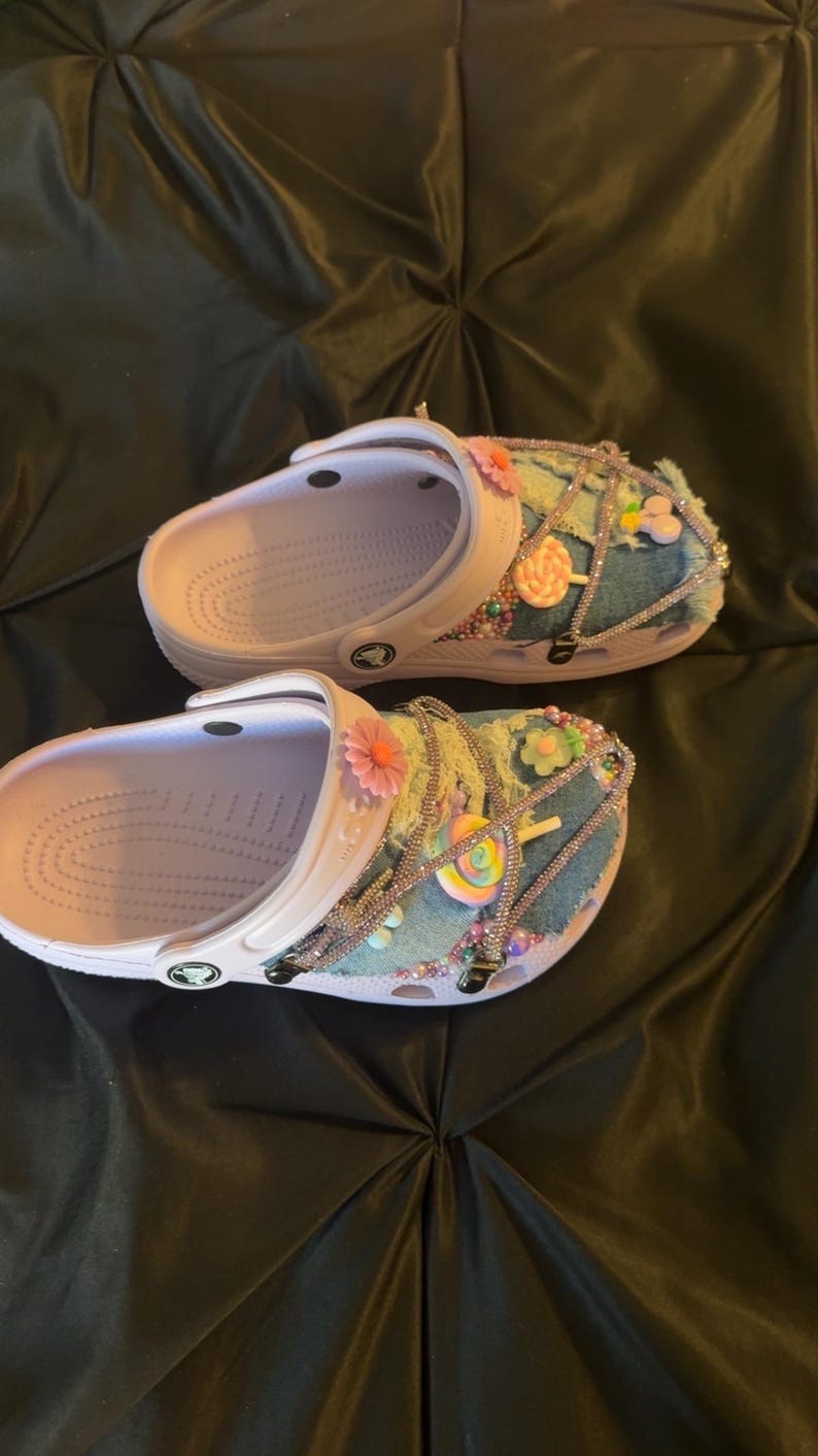 Puede incluir: Un par de zuecos blancos con plantillas de color lila claro, adornados con parches de mezclilla, pedrer&iacute;a y decoraciones florales y de caramelos de colores. Los zuecos tienen una correa y est&aacute;n sobre un fondo negro.
