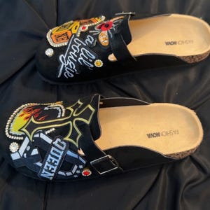 May include: Black clogs with decorative patches and embellishments. One clog features the word "Queen" and a cross design, while the other has the text "lil' bougie". Both have cork soles and are from Fashion Nova.