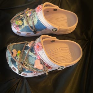 Puede incluir: Dos pares de zapatos Crocs rosas y blancos, personalizados con parches de mezclilla, flores rosas, dijes en forma de coraz&oacute;n y detalles de pedrer&iacute;a. Los zapatos est&aacute;n decorados con varios adornos y dijes.
