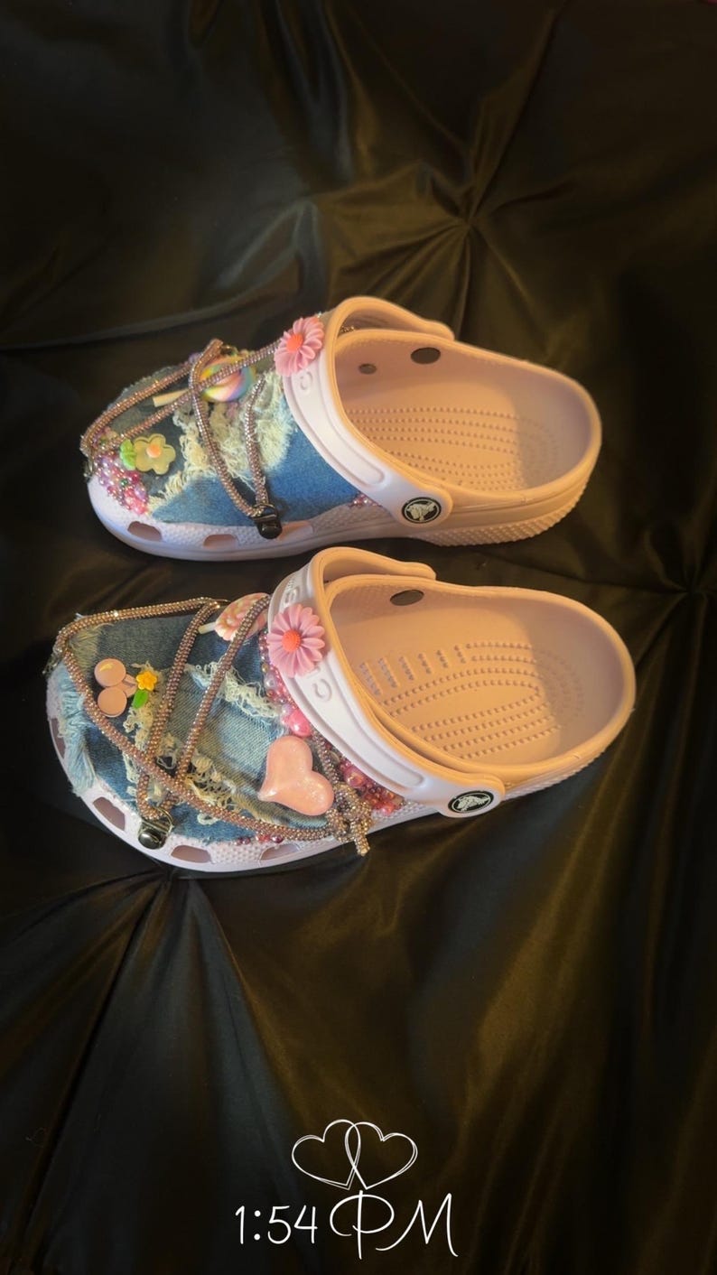 Puede incluir: Dos pares de zapatos Crocs rosas y blancos, personalizados con parches de mezclilla, flores rosas, dijes en forma de coraz&oacute;n y detalles de pedrer&iacute;a. Los zapatos est&aacute;n decorados con varios adornos y dijes.