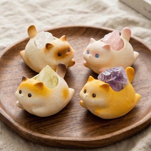 Könnte beinhalten: Vier Keramik-Katzenfiguren, die jeweils einen anderen Kristall halten. Die Katzen sind in Creme-, Gelb- und Brauntönen gehalten, mit verschiedenfarbigen Kristallen. Sie stehen auf einem Holztablett und bilden eine skurrile und dekorative Präsentation.
