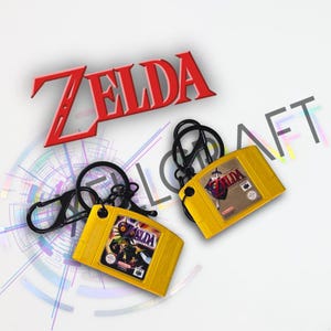 May include: Two yellow Nintendo 64 game cartridge keychains with black carabiner clips. One features The Legend of Zelda Majora's Mask label, and the other features The Legend of Zelda Ocarina of Time label. The word ZELDA is in red text above.