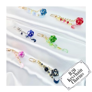 Può includere: Sei portachiavi con dadi a venti facce D20, chiusure e catene in metallo dorato. Ogni ciondolo presenta un dado blu, rosso, verde, viola o nero, arricchito da piccole perline e un pendente a forma di mezzaluna. Il testo nell'angolo recita D20 Keychain Charms.