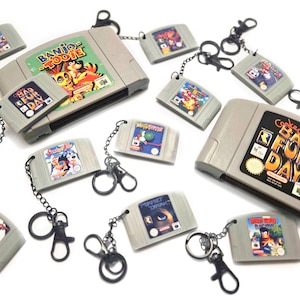 Op de afbeelding: Een verzameling miniatuur sleutelhangers van grijs plastic in de vorm van videospelcartridges met diverse titels zoals Banjo-Tooie, Conker's Bad Fur Day, Perfect Dark en Star Wars Episode I, elk voorzien van een zwarte metalen ring en karabijnhaak.
