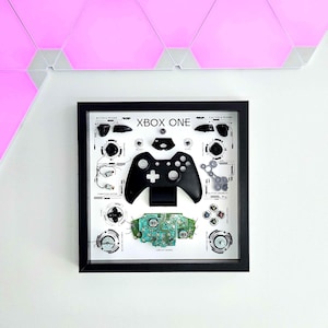 May include: Framed artwork showing the components of an Xbox One controller. A black controller is the focal point, with its parts arranged around it. The text "XBOX ONE" is displayed at the top of the frame. The frame is black.