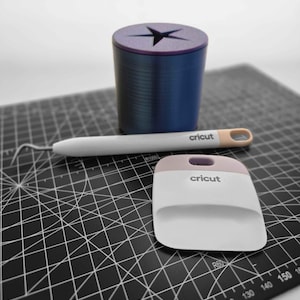 May include: A blue and purple tool holder with a star-shaped opening, a white and tan weeding tool, and a white and pink Cricut scraper sit on a black cutting mat. The cutting mat has white grid lines and measurement markings.