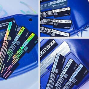 Puede incluir: Una consola de juegos portátil azul con varias etiquetas adhesivas de repuesto negras. Las etiquetas muestran el texto MODEL NO. AGB-001, YOUR TEXT HERE y el número de serie AH17103414 en letras blancas o holográficas sobre fondo oscuro.
