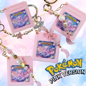 Op de afbeelding: Drie roze Game Boy-cartridge sleutelhangers met Pokémon Pink Version-labels met Mew. Elke sleutelhanger bevat goudkleurige hardware, witte en roze kralen en bedels in de vorm van sterren, manen en strikken. De tekst luidt Pokémon Pink Version.