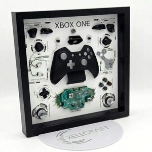 May include: A black framed shadow box display featuring a disassembled Xbox One controller. The white backing shows the controller components, including the shell, circuit board, buttons, and vibration motors, with technical labels and lines.
