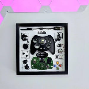 Op de afbeelding: Ingelijste kunst met een gedemonteerde Xbox Duke-controller. De zwarte controller staat centraal, omringd door zijn componenten, waaronder knoppen, printplaat en connector. De tekst "XBOX DUKE" is zichtbaar. Het kunstwerk zit in een zwart frame.
