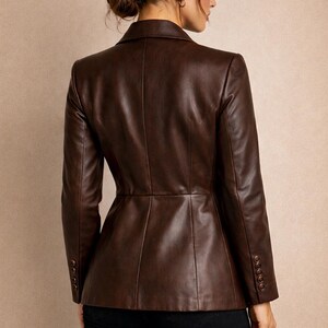 May include: A brown leather blazer with long sleeves and a tailored fit. The blazer has a button closure on the sleeves and a classic collar. The back view shows the blazer's design and stitching details. The logo "VH" is visible in the upper left corner.