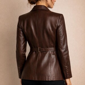 May include: A brown leather jacket with a belted waist and buttoned cuffs. The jacket has a classic blazer design and is worn over black pants. The image includes the letters "VH" in a decorative font.