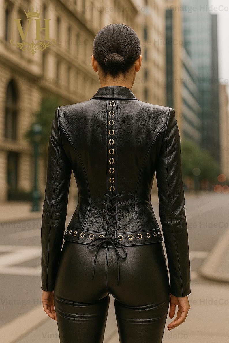 May include: A black leather jacket with a corset-style back, featuring gold-colored eyelets and a lace-up closure. The jacket has long sleeves and is paired with matching black leather pants. The image is taken outdoors.