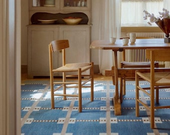 May include: A dining room scene featuring a wooden table and chairs with woven seats. A blue rug with a geometric pattern covers the floor. A cabinet with glass doors and a window with sheer curtains are in the background.