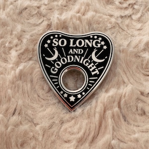 May include: A heart-shaped planchette with the words "SO LONG AND GOODNIGHT" in white lettering. The planchette is black with silver trim, featuring stars and crescent moon designs. It has a circular opening in the center.
