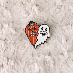 May include: A Halloween-themed enamel pin featuring a heart-shaped orange pumpkin and a white ghost. The pumpkin has a carved face with a black outline, and the ghost has a friendly expression. The pin is set against a fluffy, off-white background.