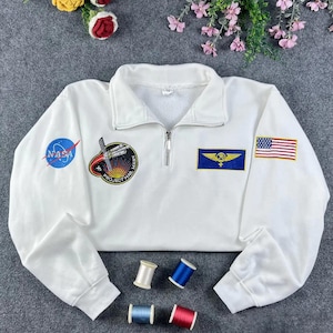 以下が含まれることがあります： NASAをテーマにした白いスウェットシャツ。クォータージップの襟付き。NASAのロゴ、アメリカ国旗、"Project Hail Mary"のパッチなどの刺繍が施されています。長袖のスウェットシャツです。