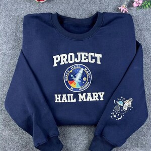 May include: A navy blue sweatshirt with the text "PROJECT HAIL MARY" in white. A circular emblem displays a rocket and the words "GRACE HAIL MARY ИПОХИНА". The sleeve features an embroidered astronaut and stars.