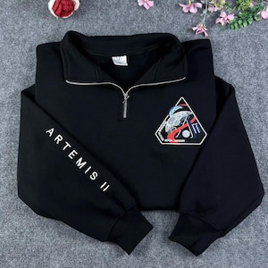 May include: Black sweatshirt with a quarter-zip collar and the text "ARTEMIS II" printed in white on the left sleeve. A triangular patch on the chest features a space-themed design with the text "KOCH HANSEN".